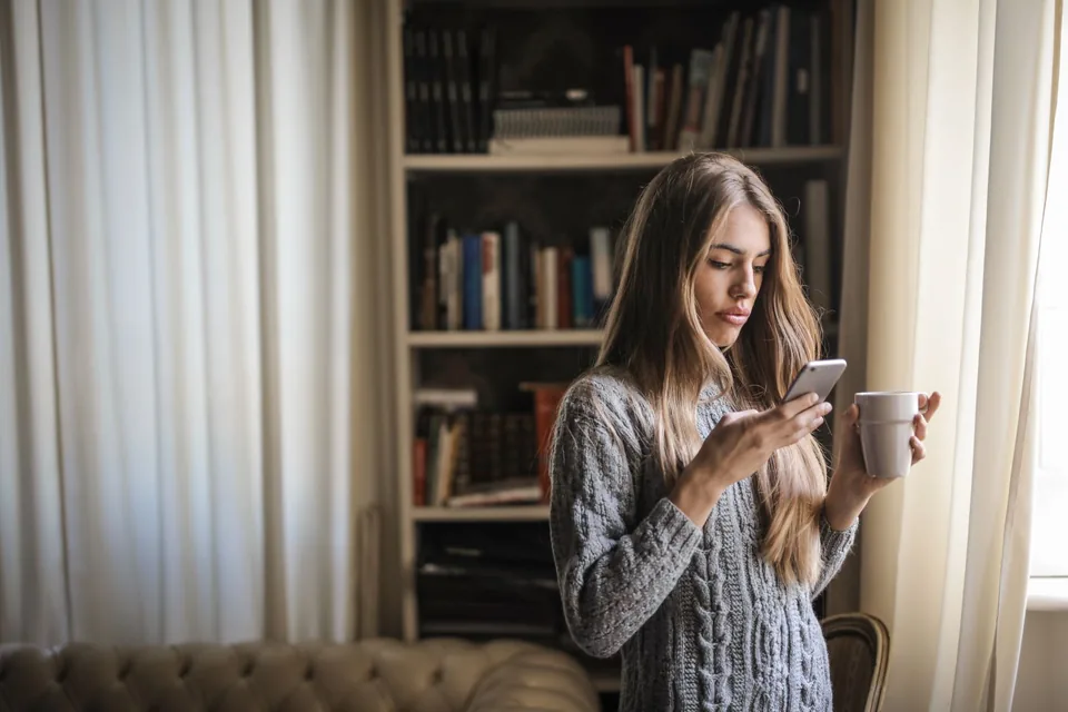 Nő reggeli kávéval otthon nézi a telefonját